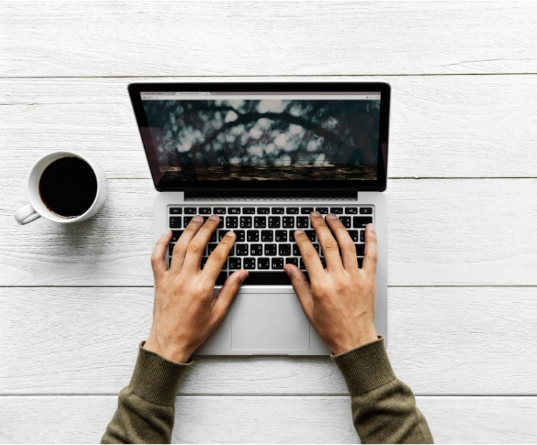 Plano aéreo das mãos de uma pessoa digitando em um notebook, com uma caneca de café ao lado